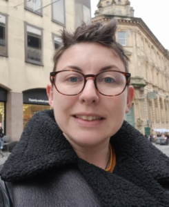 Portrait of January Star Student Charlie Taylor smiling outdoors on a city street, wearing round glasses and a black shearling jacket, with historic buildings in the background.