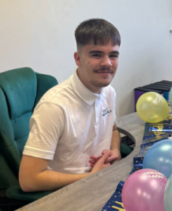 Alfie Weston, May Star Student at Accountancy Learning, smiling while seated at a desk decorated with colourful balloons in the office, wearing a white branded polo shirt.