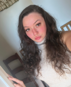 Young woman with long dark curly hair and a nose piercing wearing a sleeveless cream turtleneck top, posing indoors in natural sunlight and looking directly at the camera.